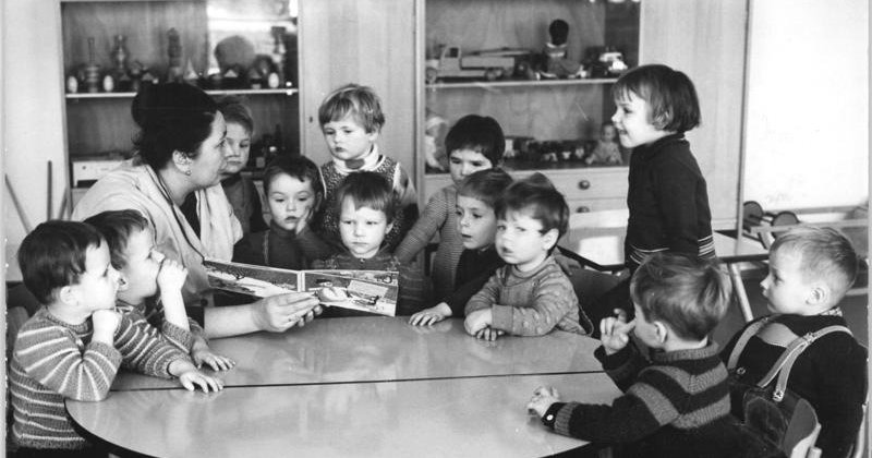 Schwarzweißfoto von 1966: Eine Erzieherin liest einer Gruppe von Kindergartenkindern an einem runden Tisch aus einem Bilderbuch vor, dahinter ein Regal mit Spielzeuglastern.