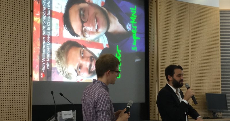 Zwei Männer mit Mikrofonen stehen vor einer großen Leinwand, auf der die Titelfolie eines Snapchat-Workshops mit zwei Selfies und grünen Kritzeleien zu sehen ist.