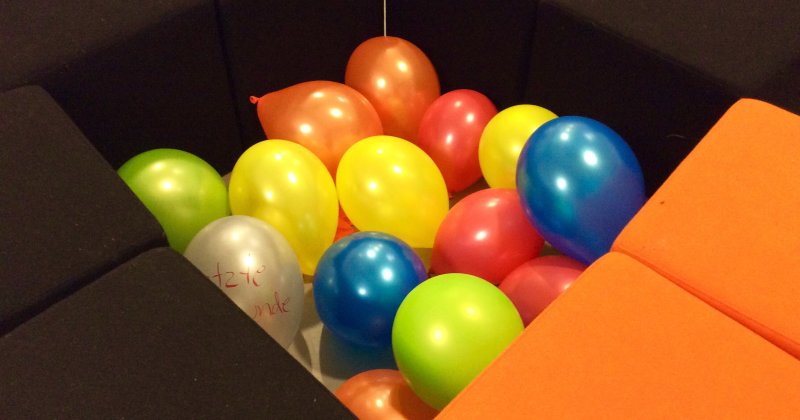 Bunte Luftballons liegen in einer Grube aus schwarzen und orangefarbenen Sitzwürfeln, auf einem weißen Ballon steht etwas Handschriftliches.